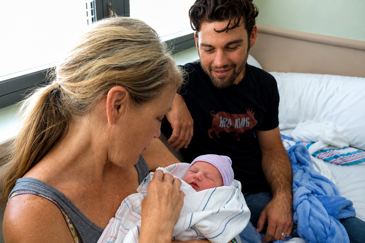 Grandmother cradling her newborn grandchild in a hospital room while the baby's father looks on with a smile, capturing a quiet, multi-generational family moment filled with love and connection.